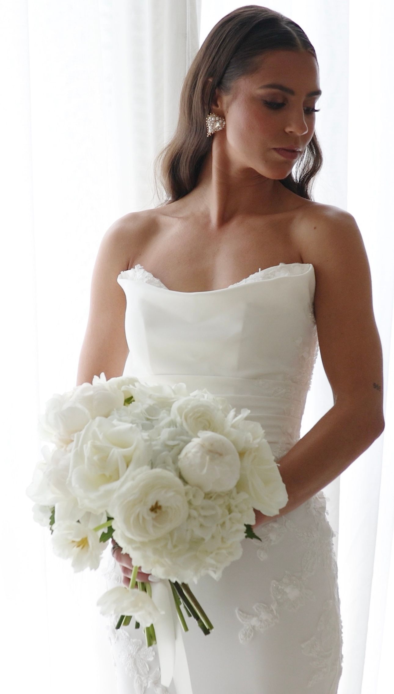 A beautiful bride in a white strapless gown holds an elegant bouquet of white peonies against a light background.