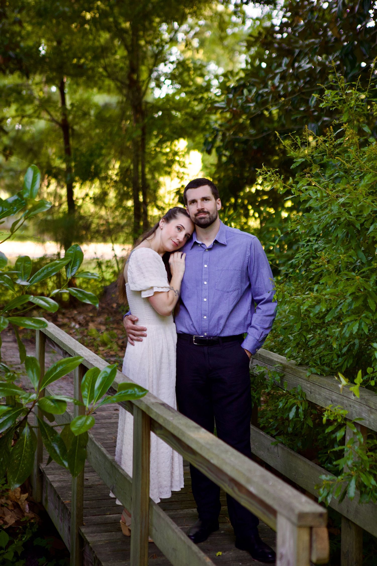 A couple stands together on a wooden footbridge surrounded by lush green foliage in a romantic outdoor setting.