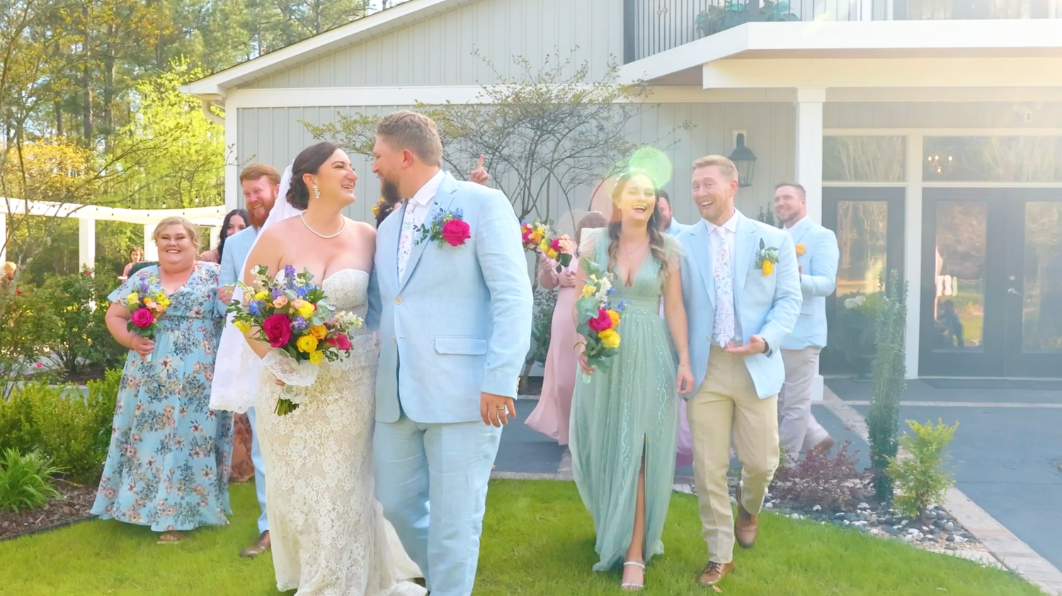 Wedding party in pastel blue and mint green attire poses together on outdoor patio with vibrant floral bouquets.