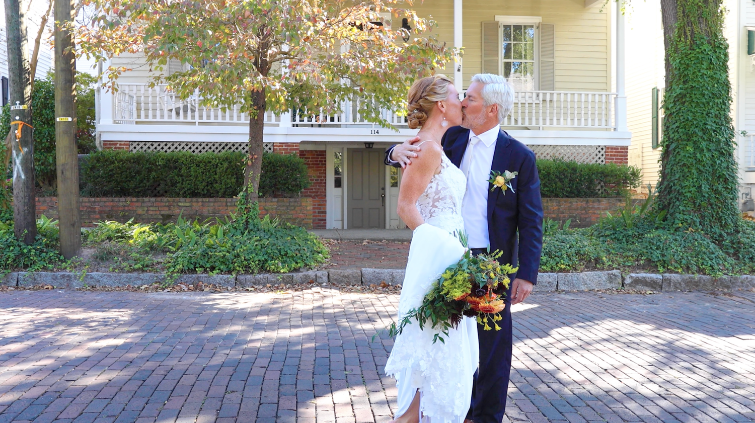 Newlyweds share an intimate moment on a brick path in front of a yellow house with white porch columns.