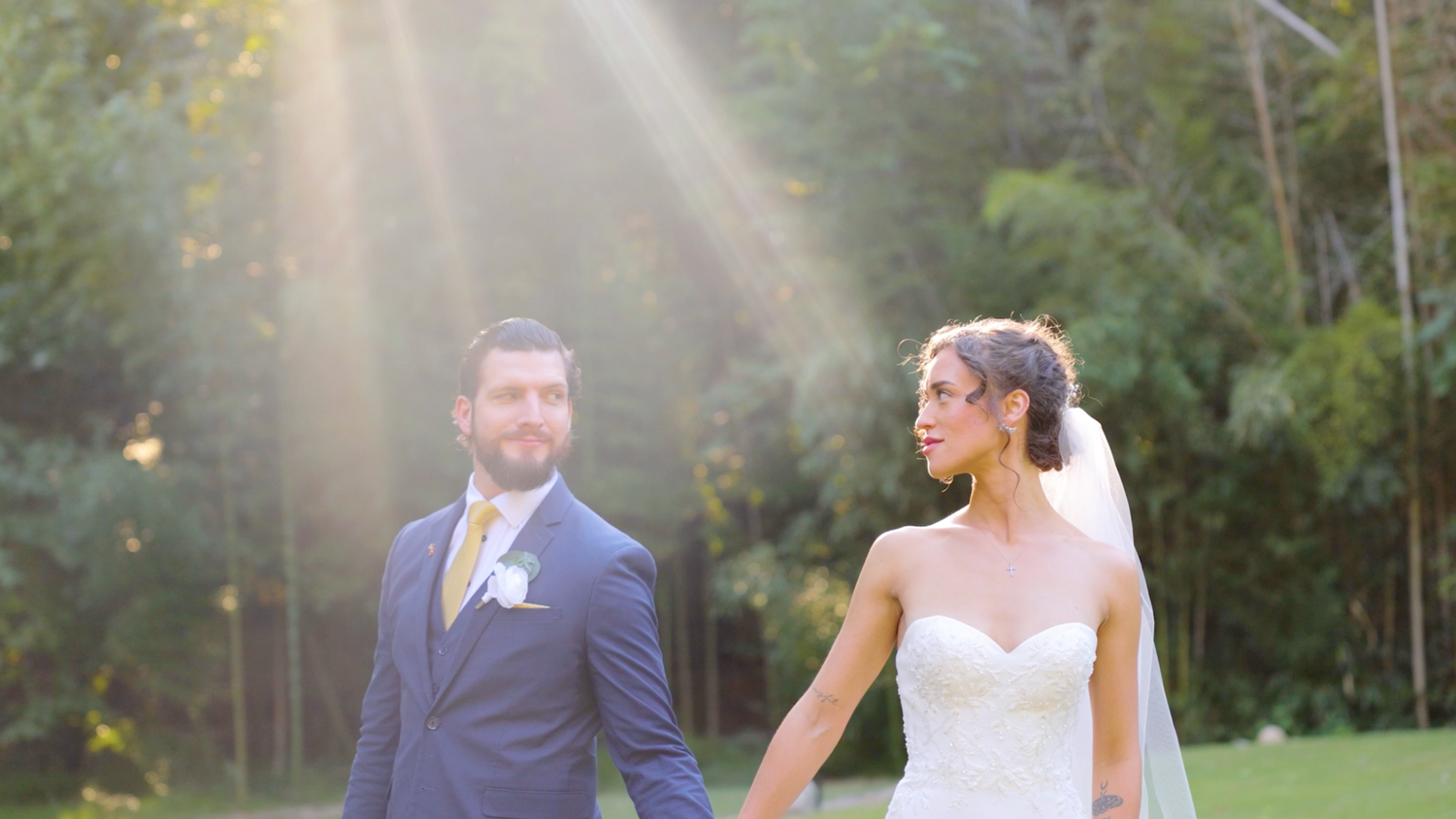 A couple holding hands walks together outdoors as sunlight streams through trees in a romantic wedding moment.