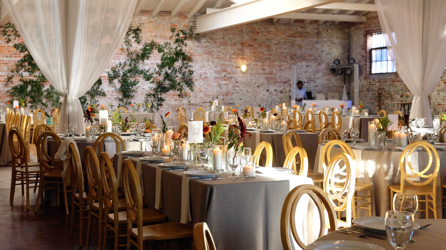 Elegant wedding reception venue with white draping, gold chairs, and floral centerpieces in a rustic brick-walled space.