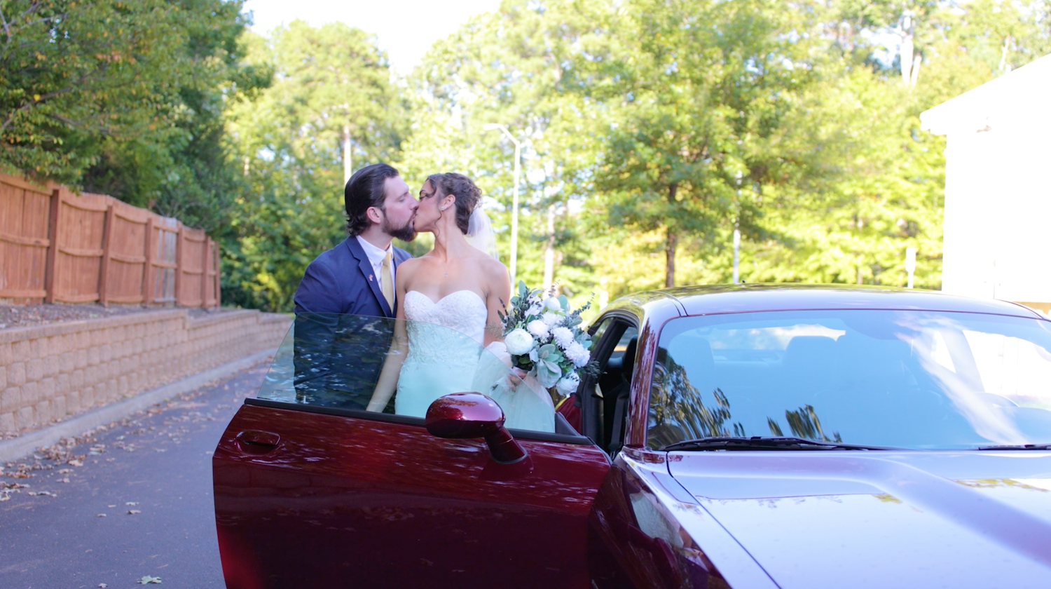 A group of people with wedding attire mingle outdoors near vehicles and trees in a sunlit setting.