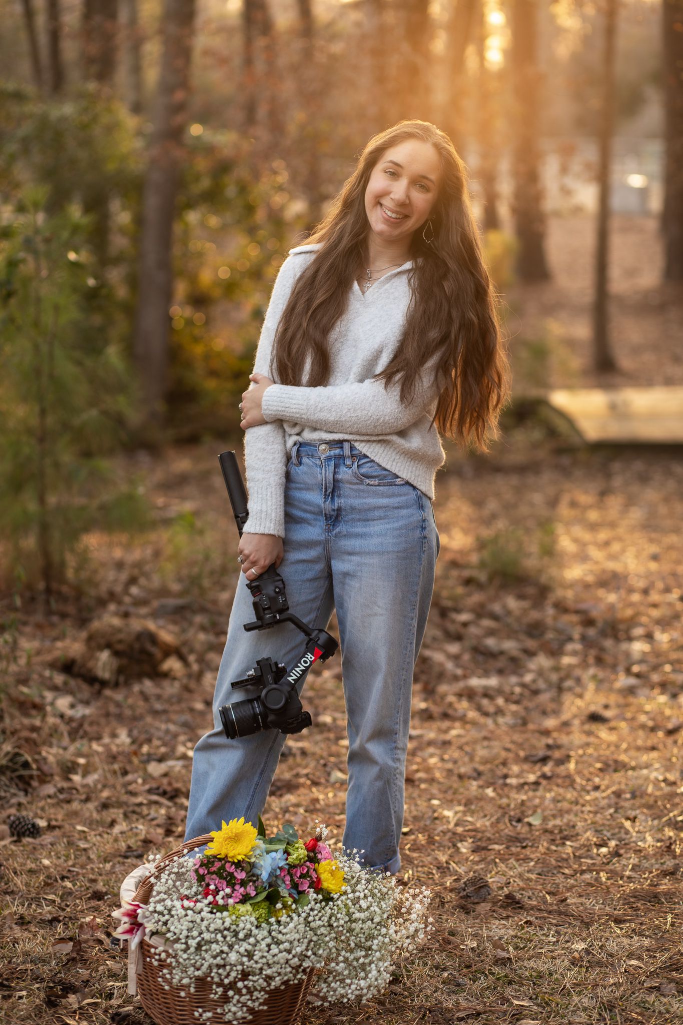 White sweater and jeans outfit with autumn forest backdrop and basket of wildflowers during golden hour sunset.