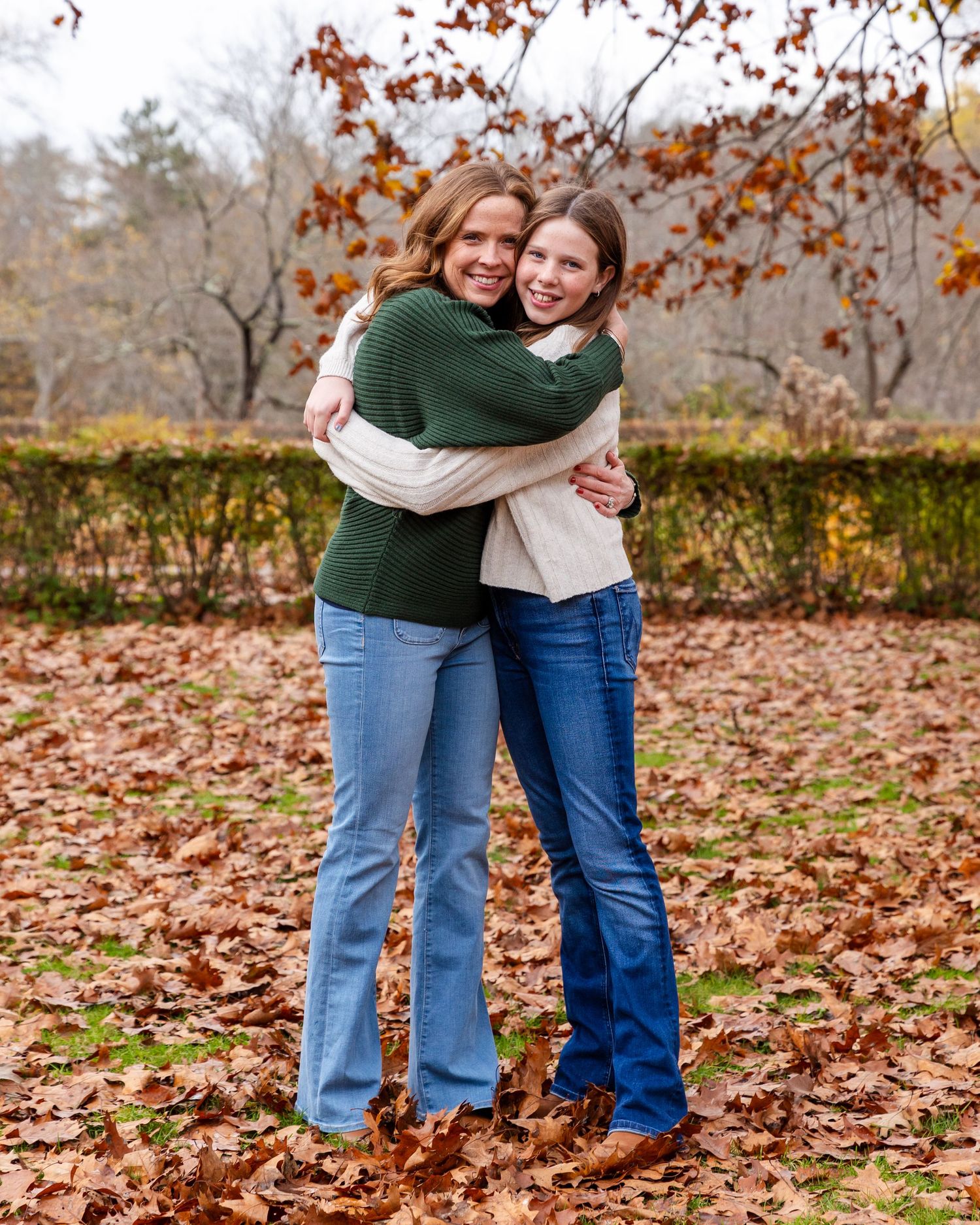 Two people wearing jeans share a warm embrace while standing on autumn leaves in front of a hedge on a fall day.