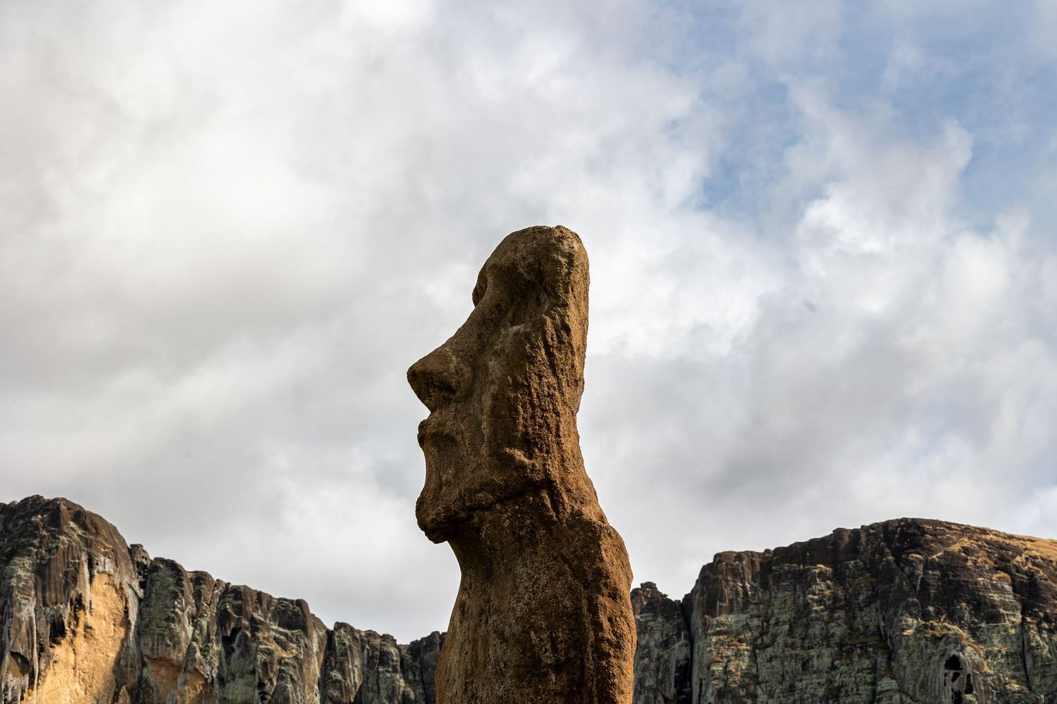 Ancient Easter Island Moai statue stands tall against cloudy sky with rocky cliffs in background.