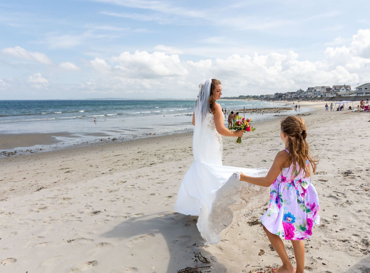Someone in white walking along a sandy beach on a sunny day with rolling waves and coastal homes in the background.