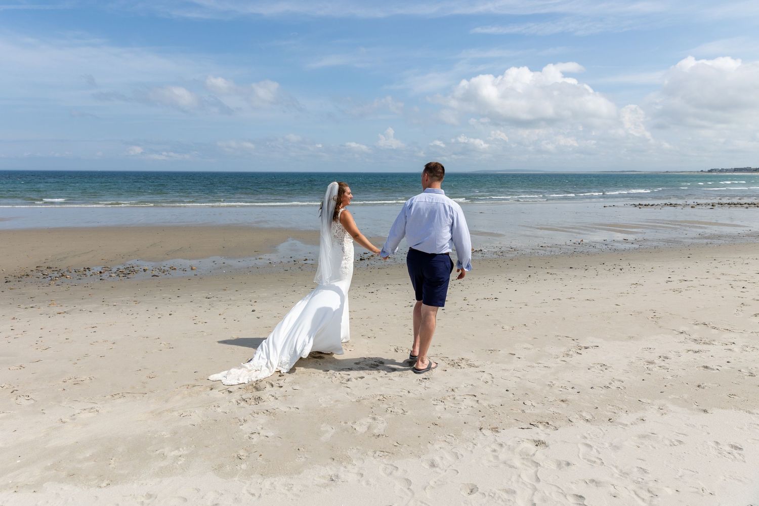 Two people walk along a white sandy beach with blue ocean waves and clear sky in the backdrop highlighting a romantic moment.