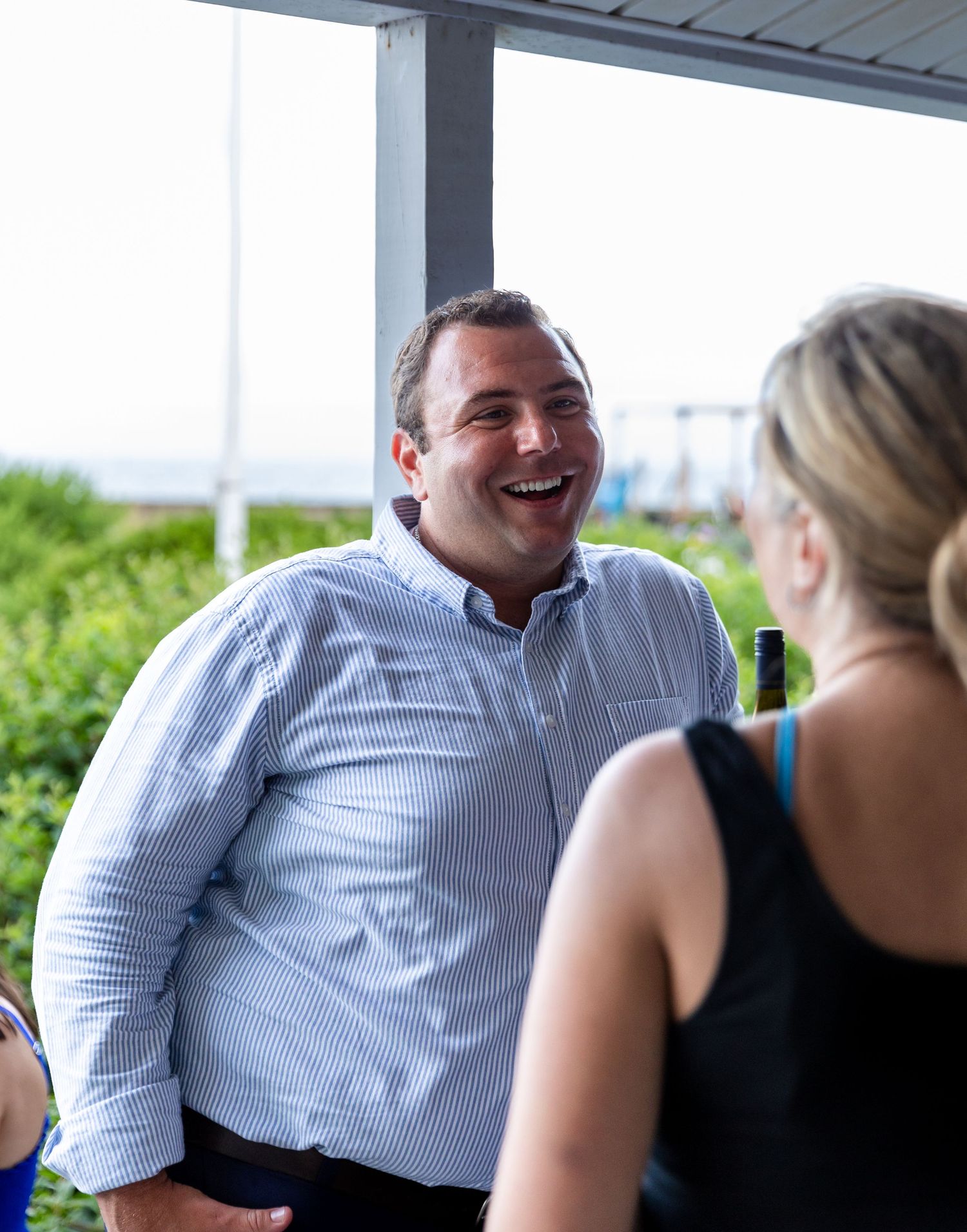 A person in a light blue striped button-down shirt laughs during an outdoor conversation on a covered patio.
