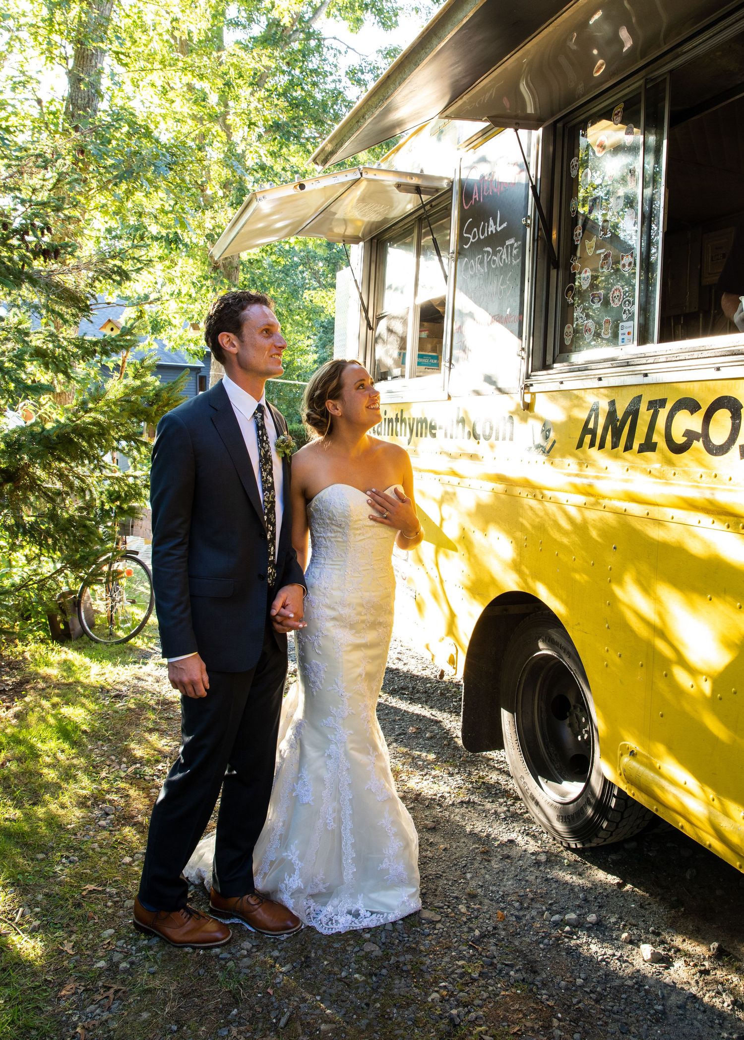 A couple in formal wedding attire stands next to a bright yellow vintage food truck called 'Amigo' in a wooded outdoor setting.