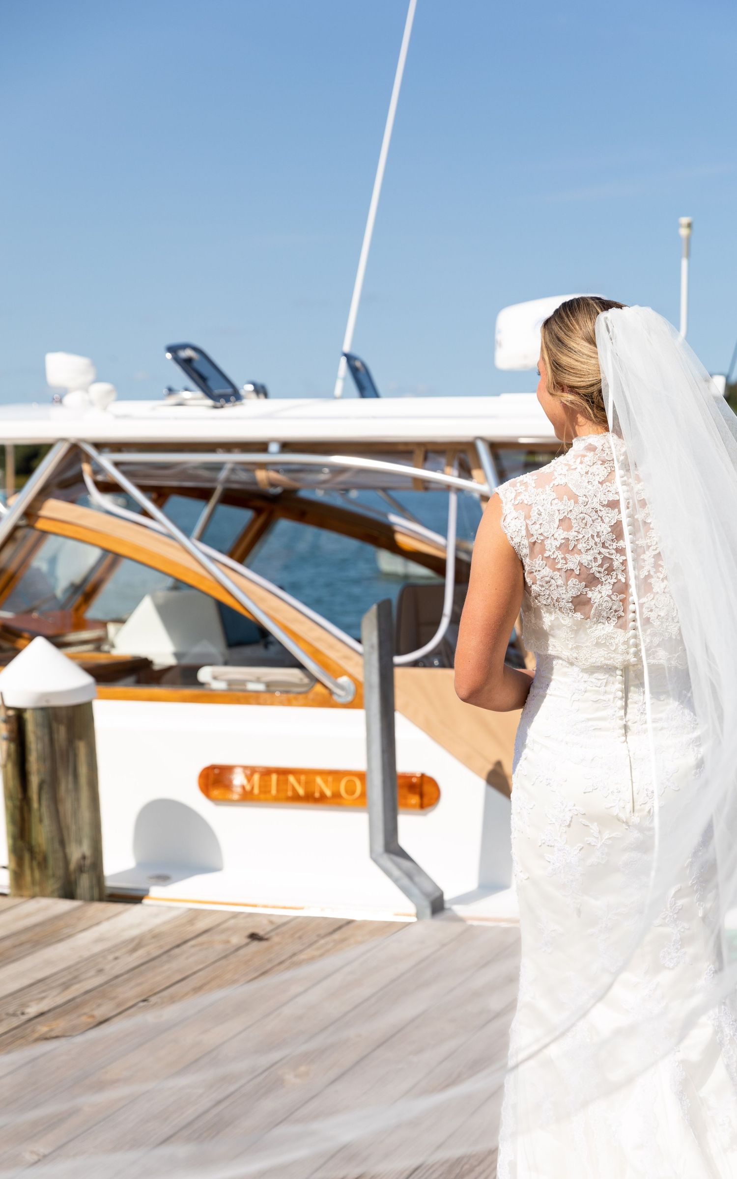 A bride in a white lace wedding dress and veil stands on a wooden boat deck overlooking the ocean.