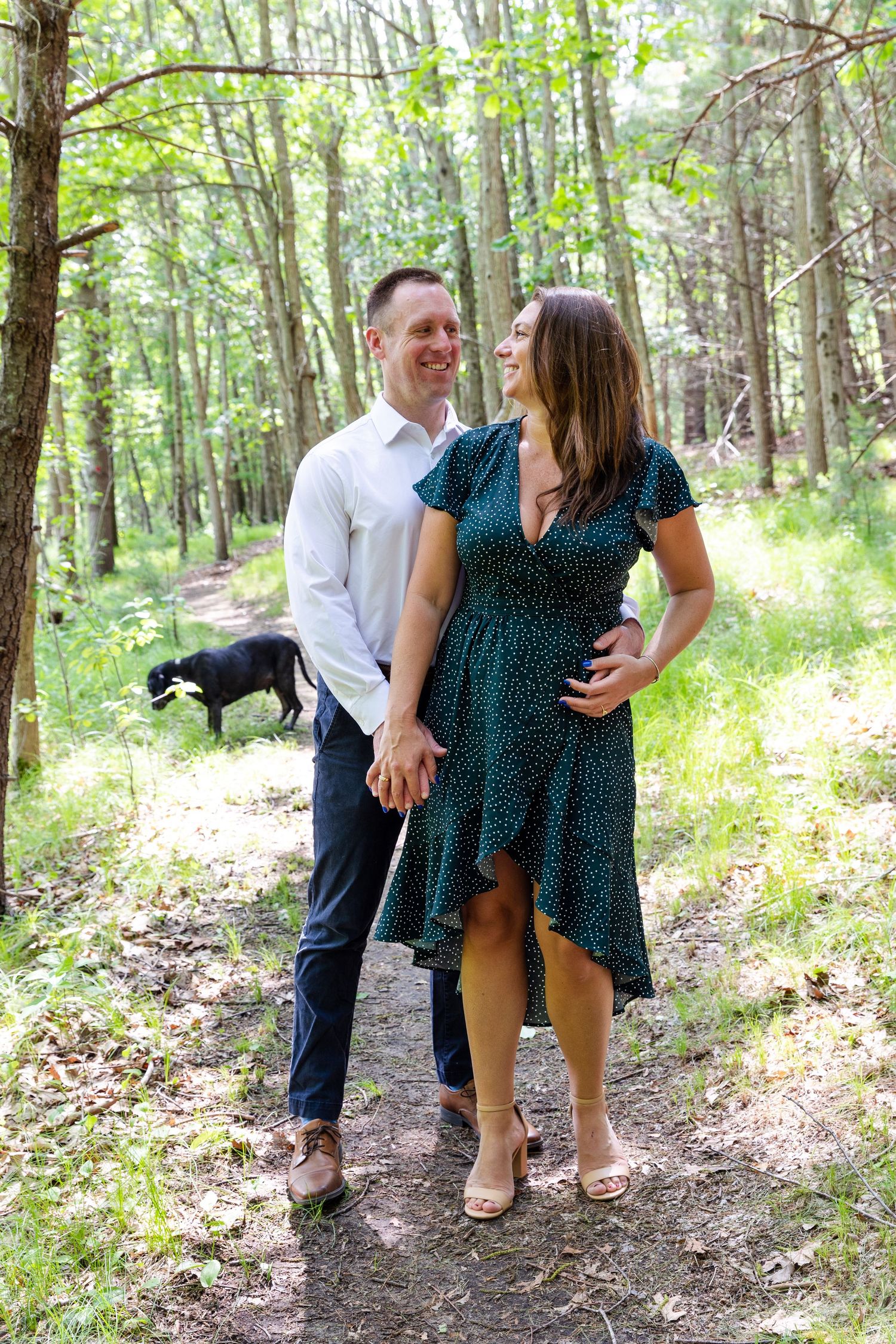 A couple shares a joyful moment with their dog while walking through a sunlit wooded area in summertime.