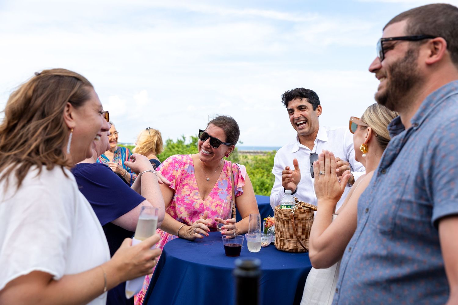 A cheerful group gathers outdoors around a blue table, enjoying drinks and conversation on a sunny day.