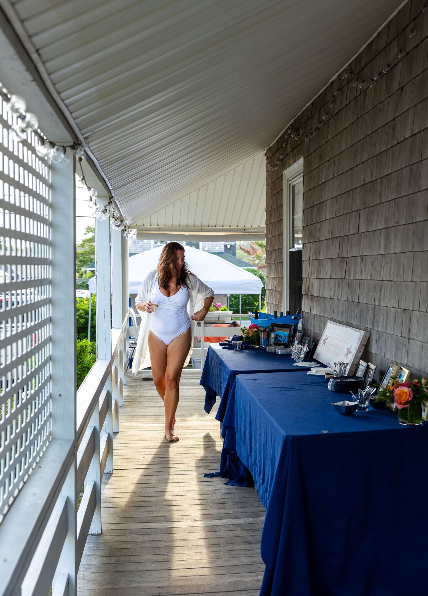 Tables with navy blue linens and refreshments line a covered wooden deck by a shingle-sided building.