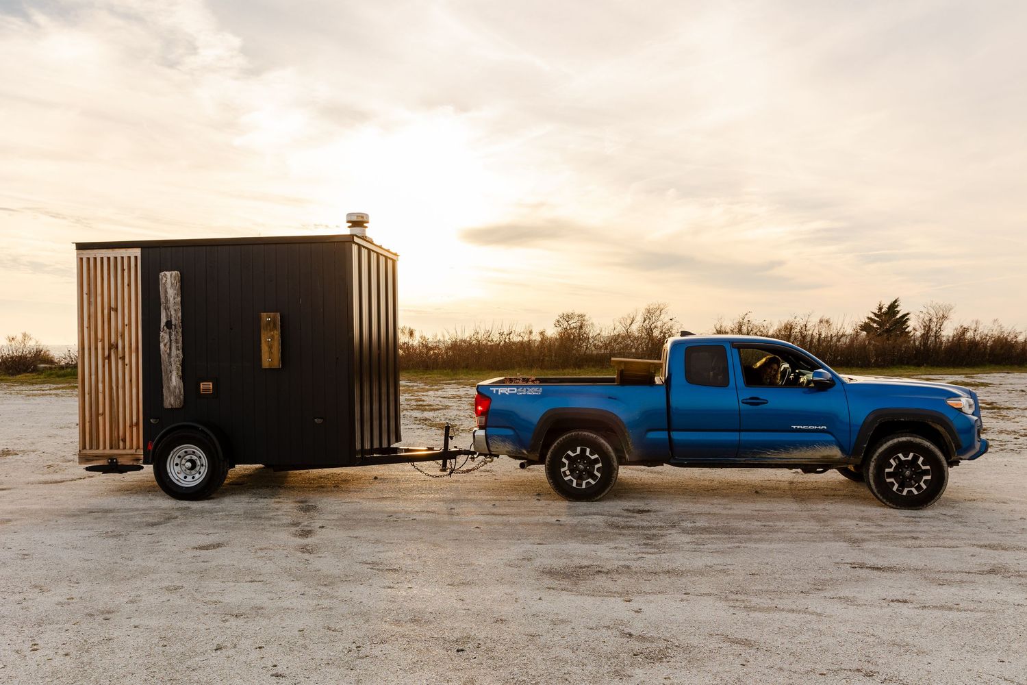 Blue pickup truck towing a wooden mobile sauna trailer at sunset on a snowy winter day.