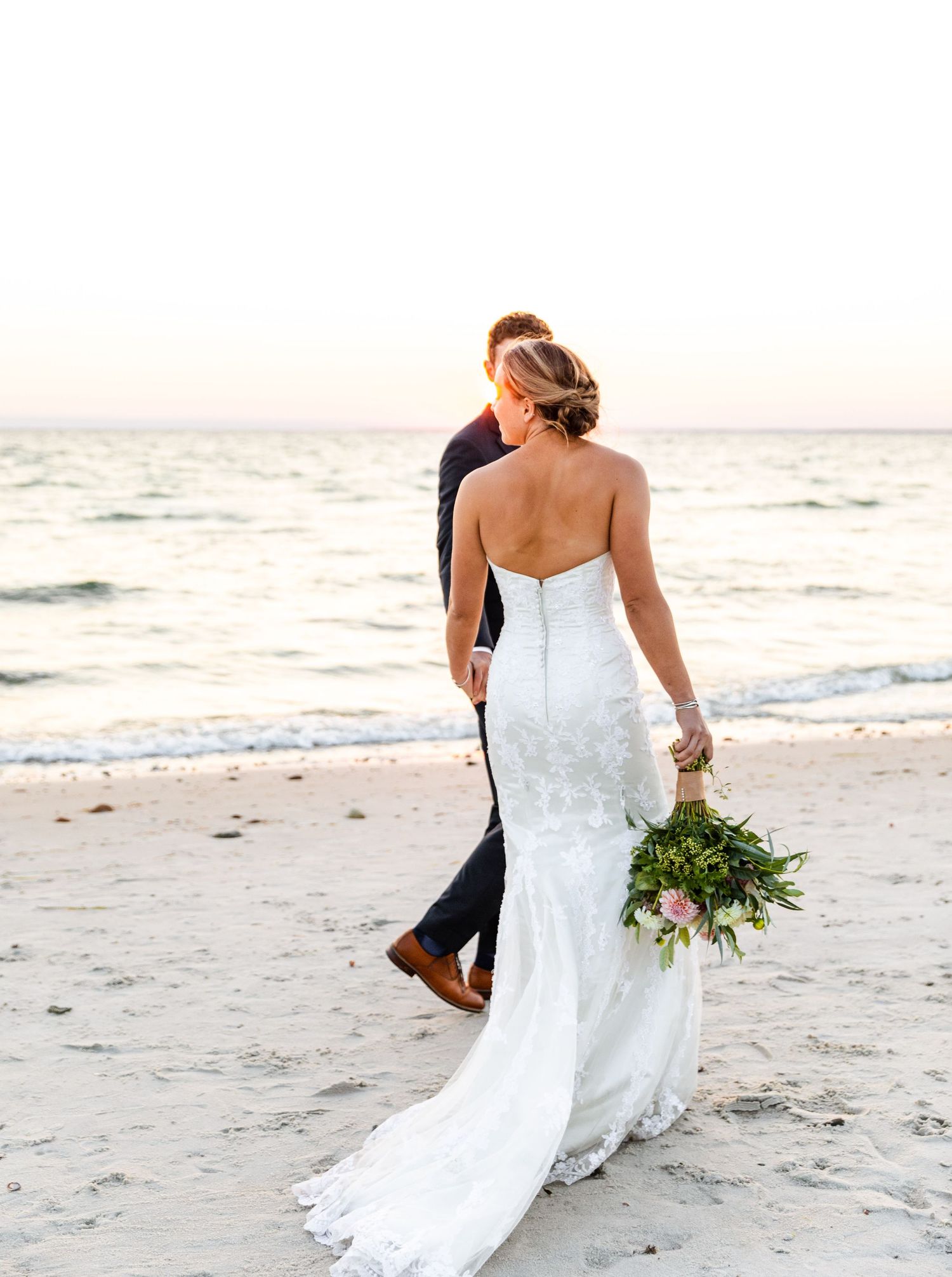 A couple walks hand in hand along a serene beach at sunset with the bride wearing a flowing white wedding dress.