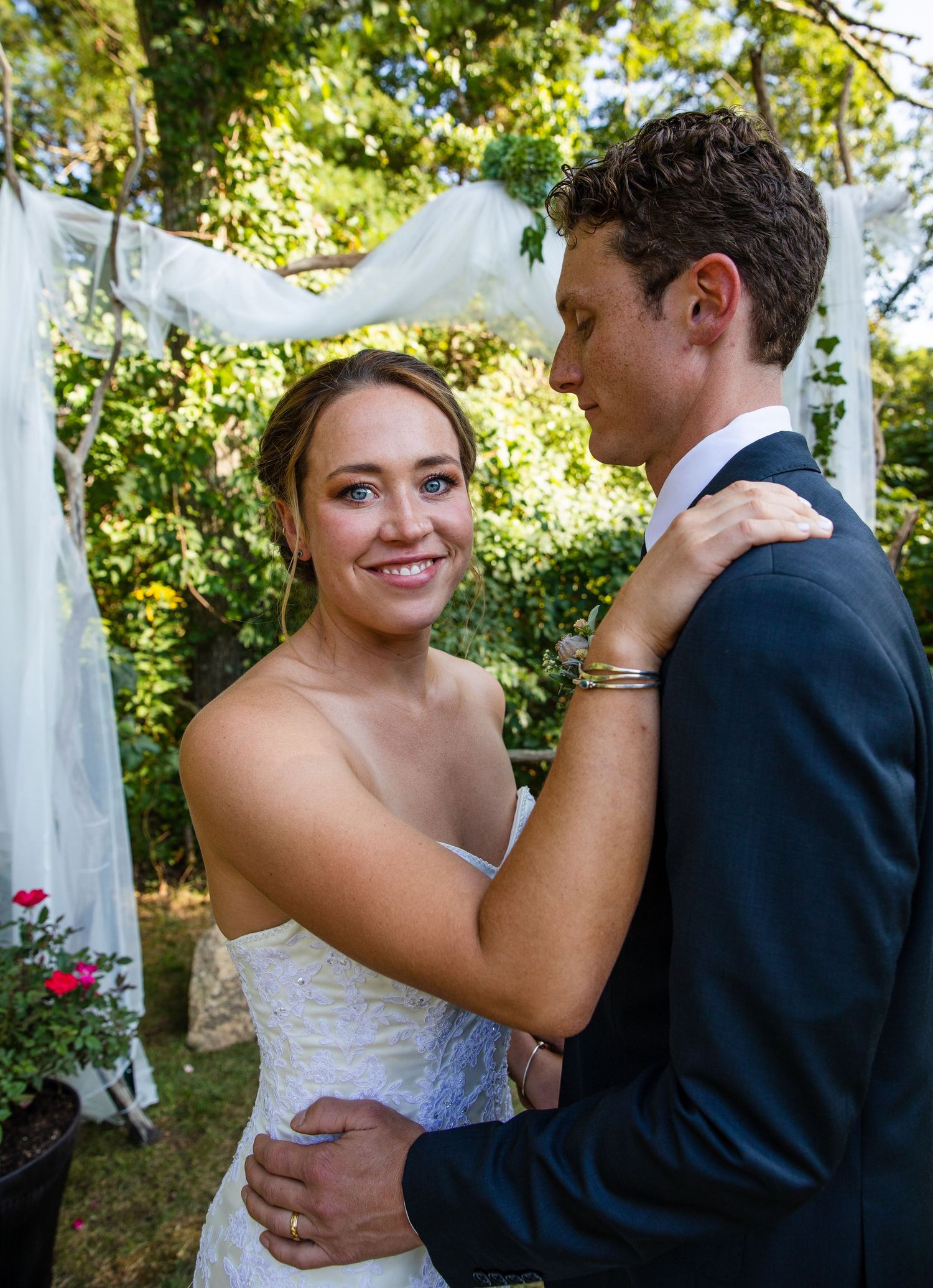 A couple in wedding attire embraces during an outdoor garden ceremony with white drapery and roses in the background.