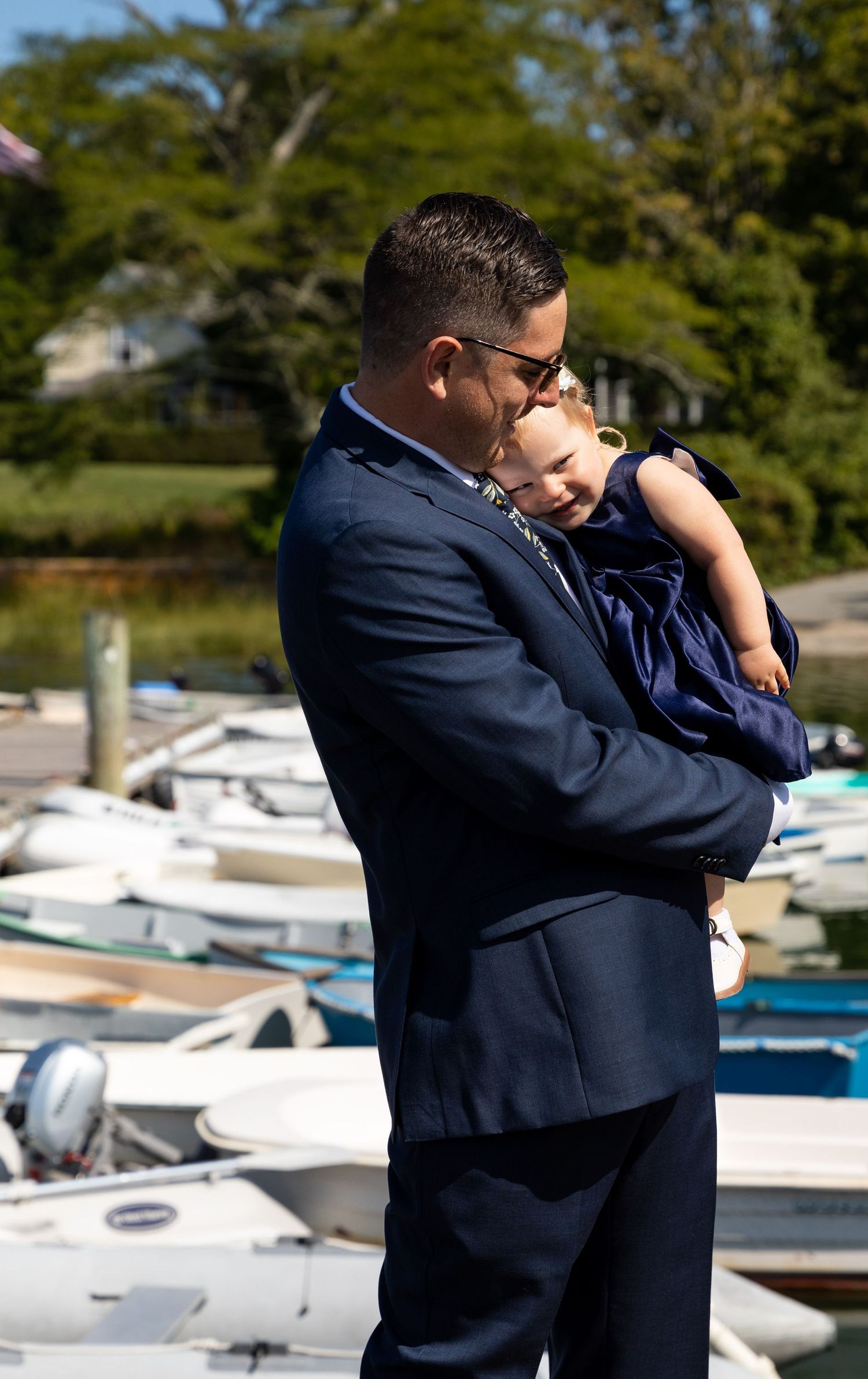 A navy-suited figure holds a sleeping baby near a marina filled with boats on a sunny day.
