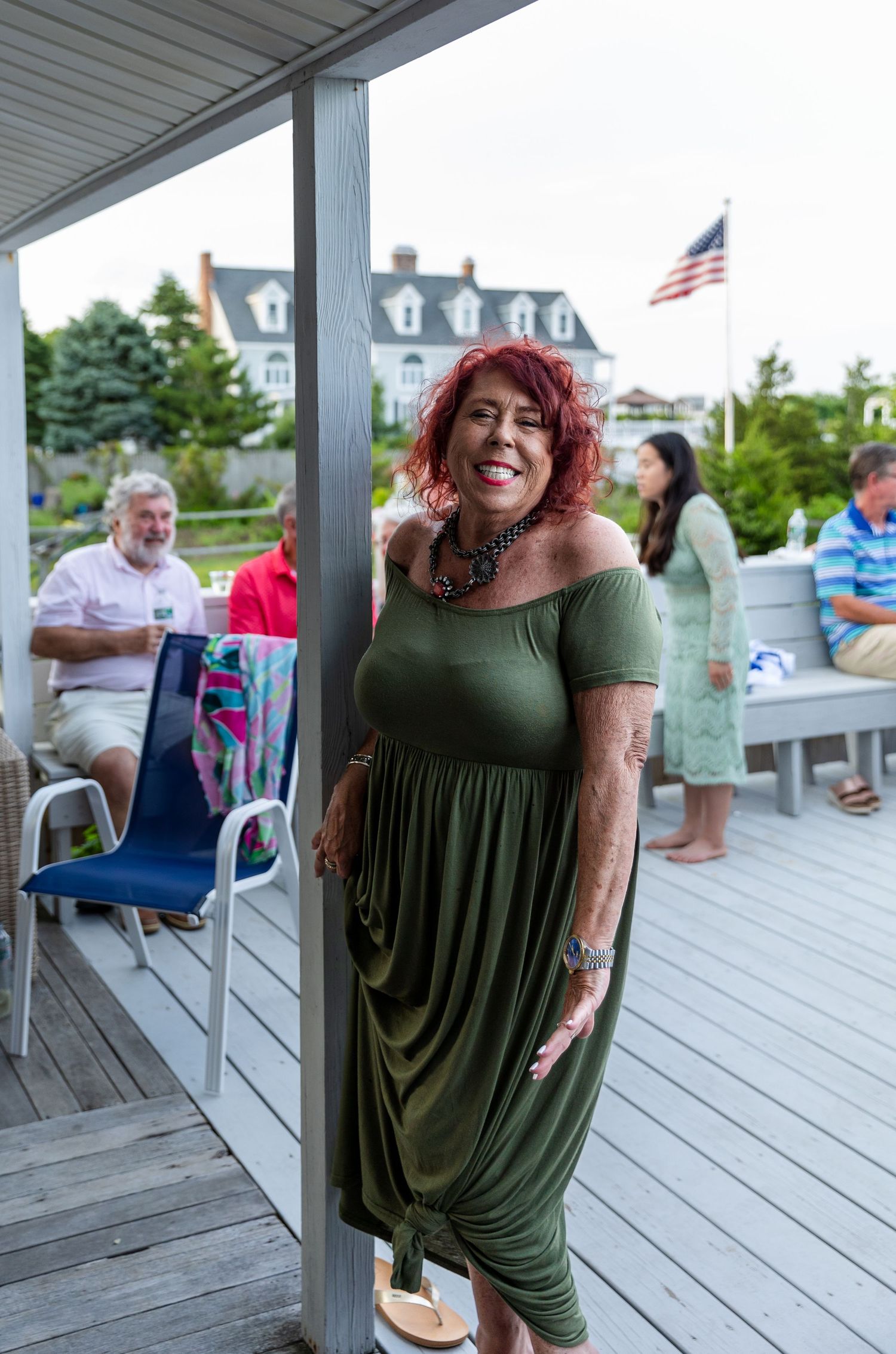 A person in a green maxi dress smiles while standing on a covered deck with other people and an American flag in view.