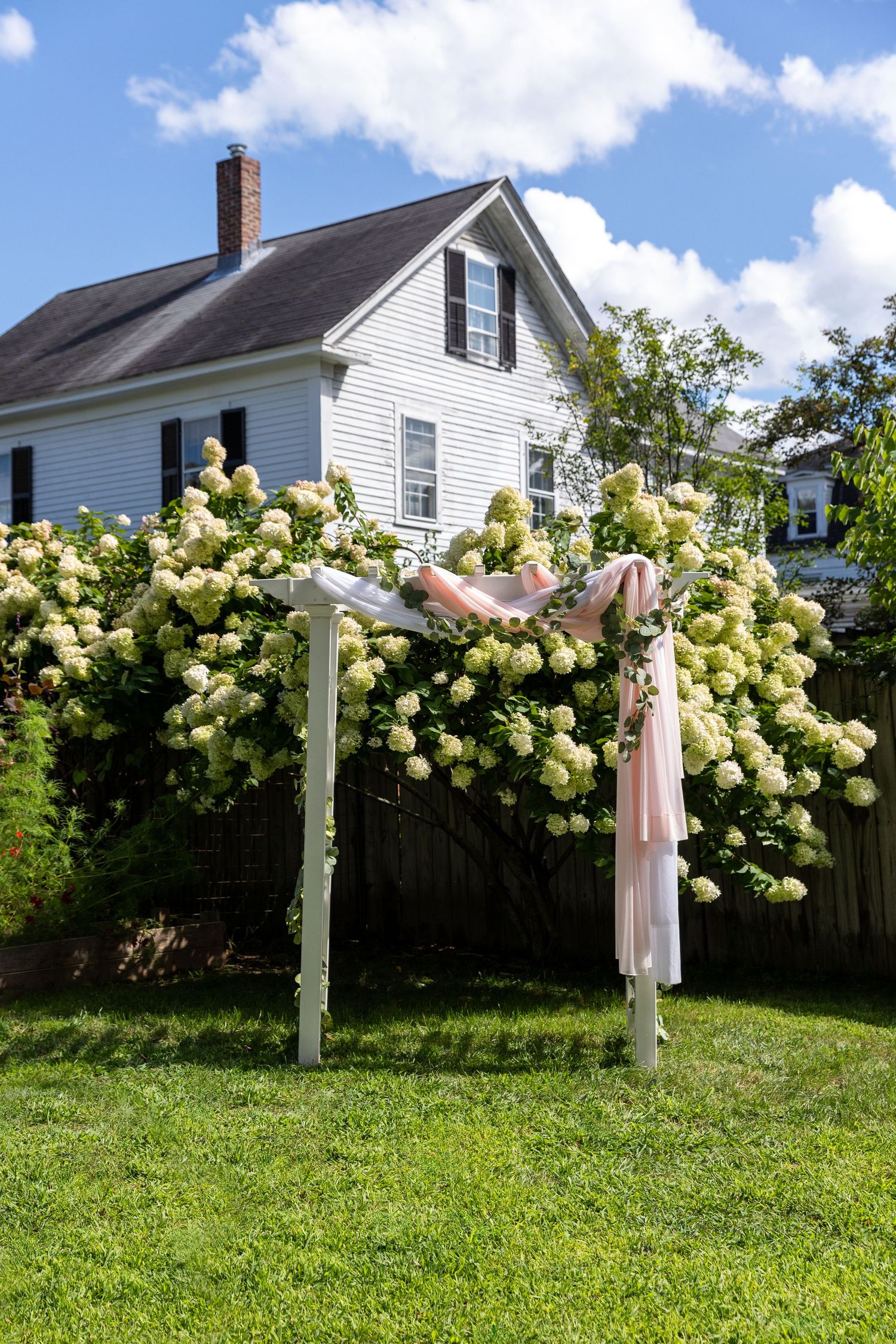 White wedding arch decorated with delicate fabric and lush hydrangea blooms stands in front of a classic white farmhouse.
