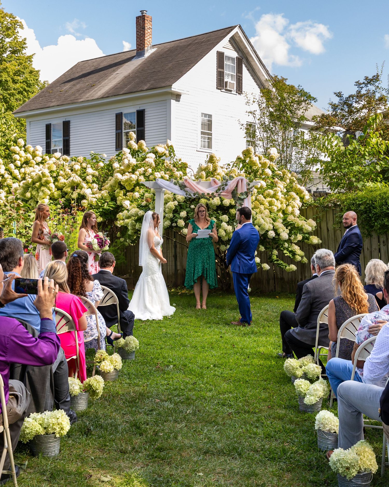 A panoramic view of an outdoor garden wedding ceremony with guests seated on white chairs and hydrangea bushes in bloom.