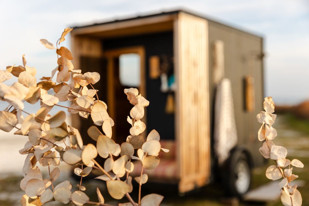 Dried floral decorations frame a wooden mobile tiny house or trailer in soft focus.