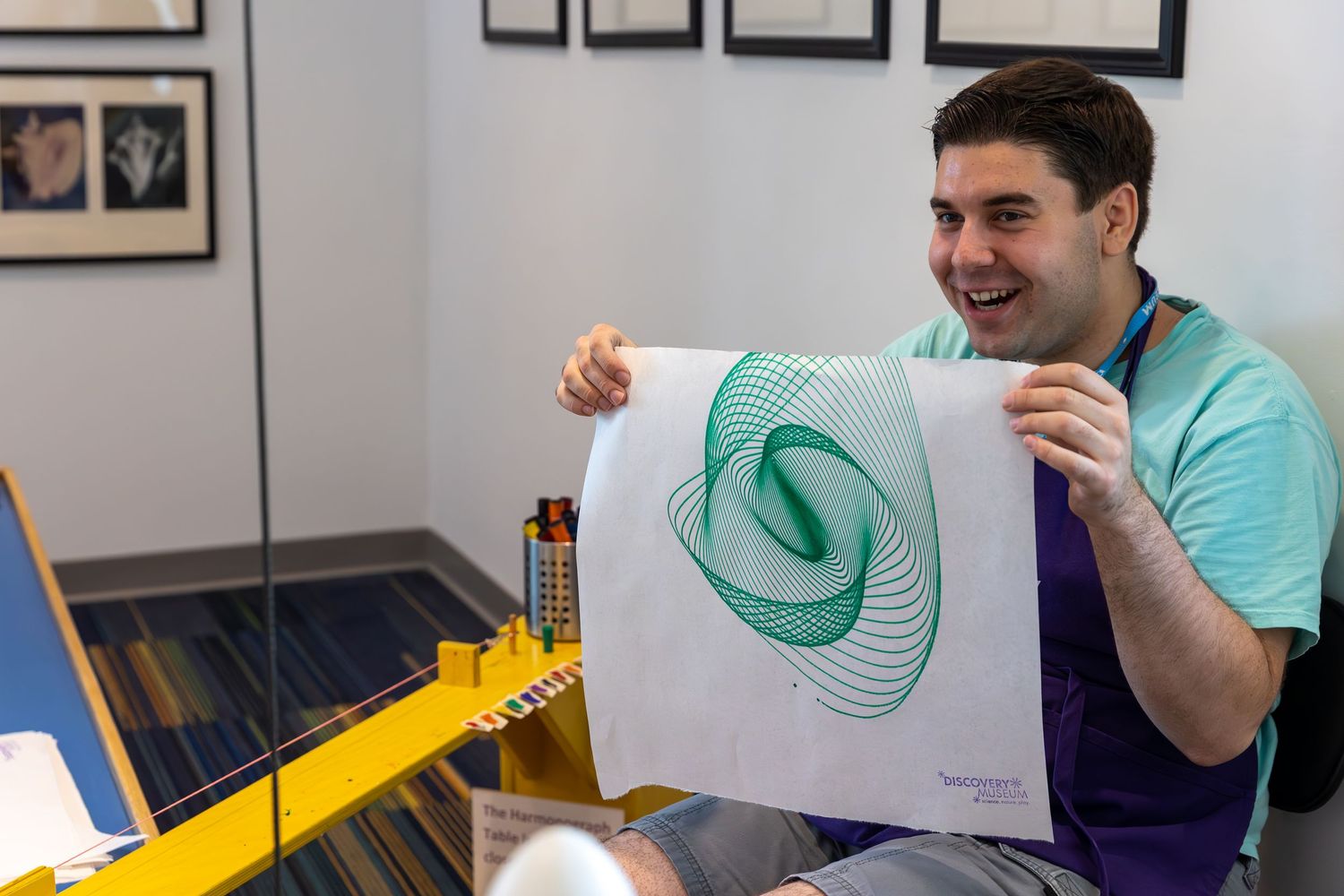 Two people laugh while sitting at a desk with framed artwork visible on the wall behind them.