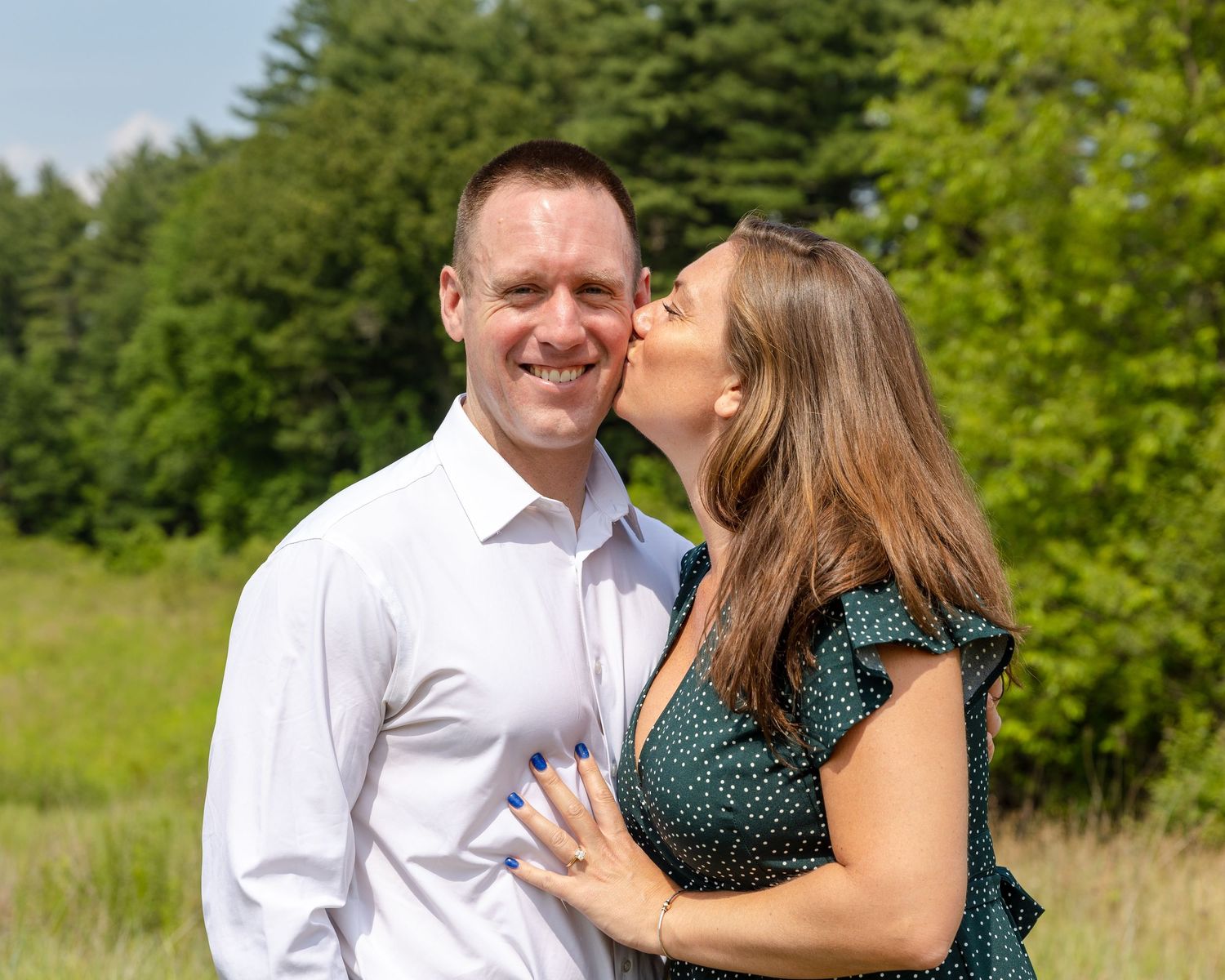 A couple in semi-formal attire embraces lovingly in a sunny meadow surrounded by lush green trees.