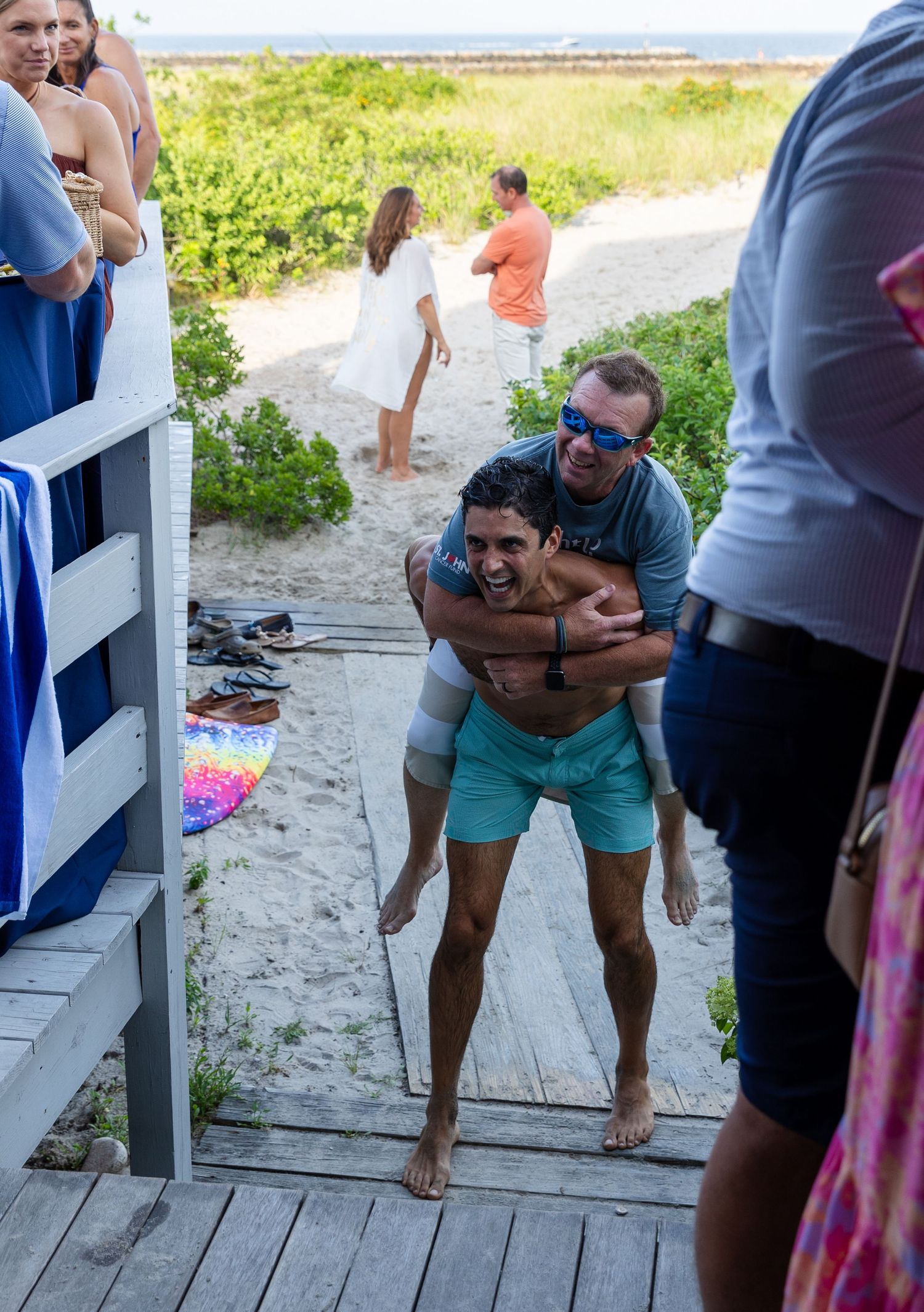 Several people in beachwear gathered on a wooden boardwalk path leading to the beach on a sunny day.