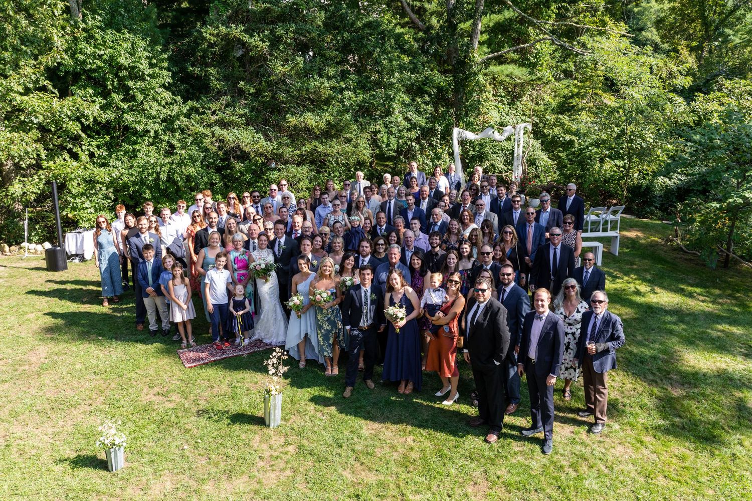 Large outdoor group photo of wedding guests gathered together on a sunny grassy lawn surrounded by trees.