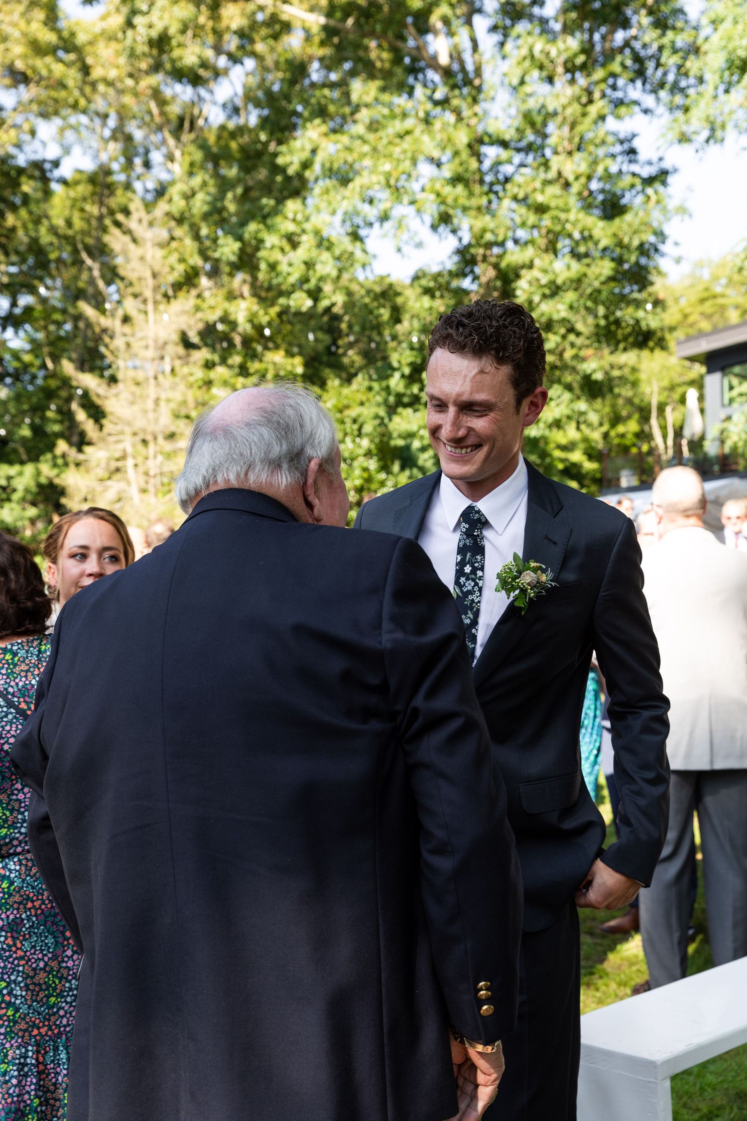 A person in a black suit greets an elderly man in formal attire at an outdoor gathering.
