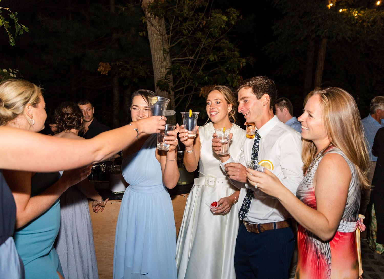 Wedding guests in light blue dresses raise their glasses together for a celebratory toast at outdoor evening reception.