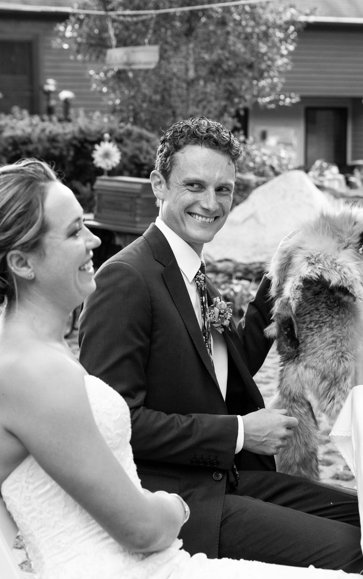 Black and white photo showing a happy couple sharing a heartwarming moment with a furry pet during their outdoor celebration.