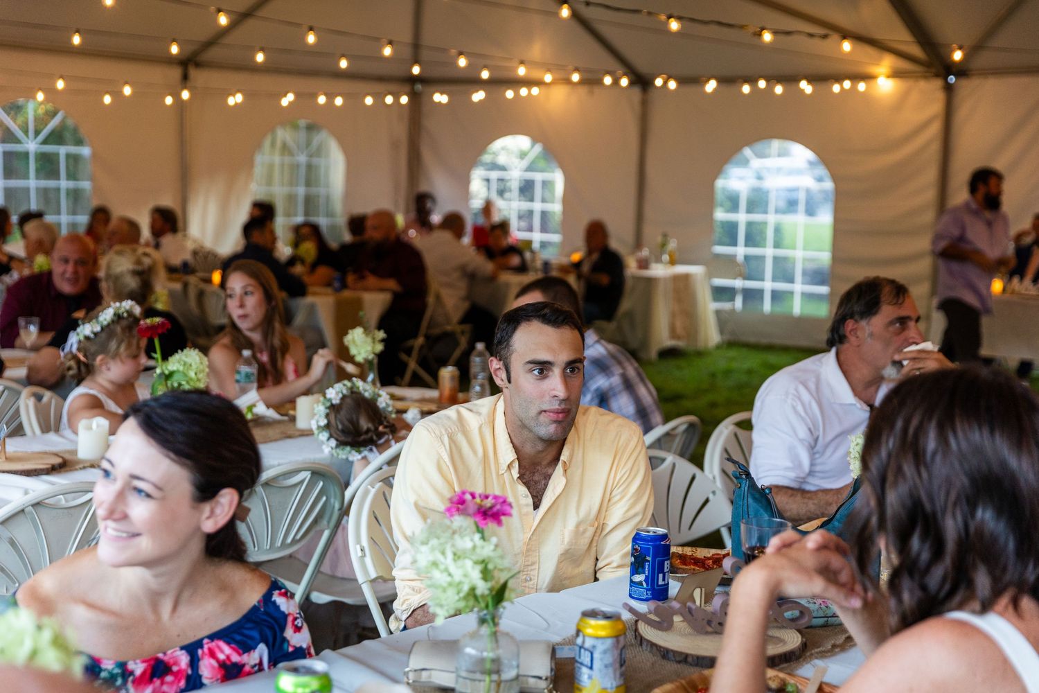 Wedding reception guests sit at round tables under a white tent decorated with string lights and floral centerpieces.