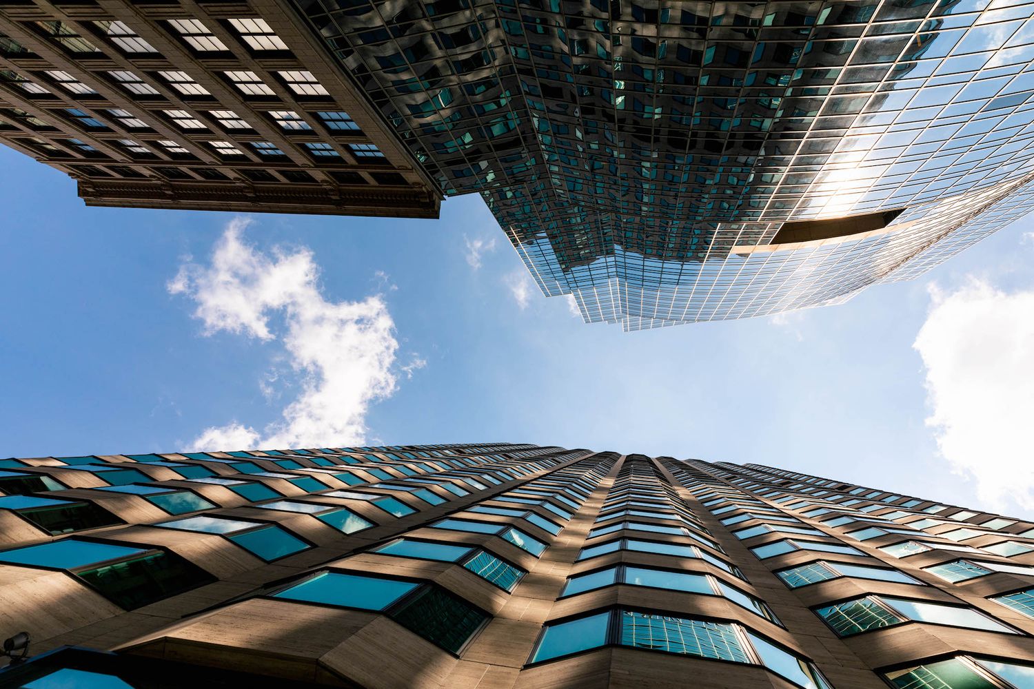 Looking up at modern skyscraper with geometric blue glass windows and angular architecture against blue sky.
