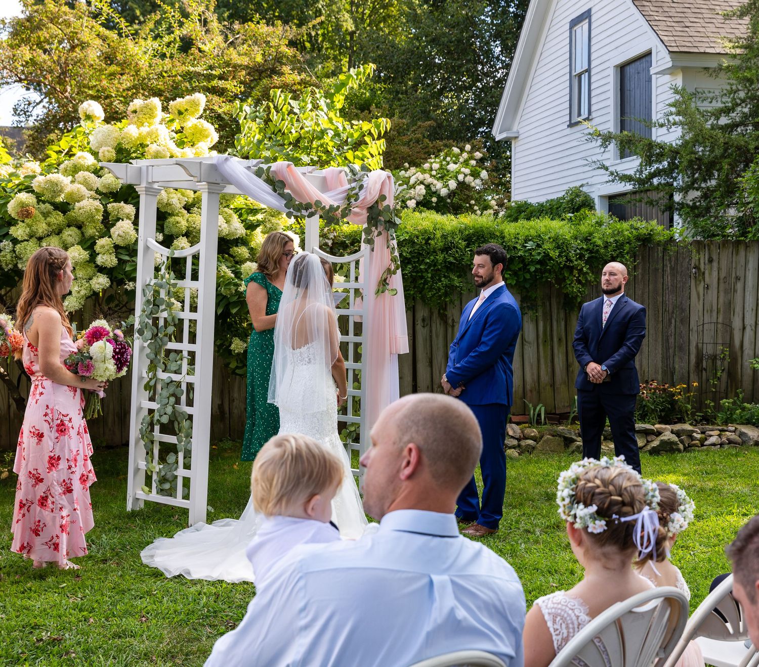 A wedding ceremony takes place under a white garden arch decorated with flowers in a backyard setting.