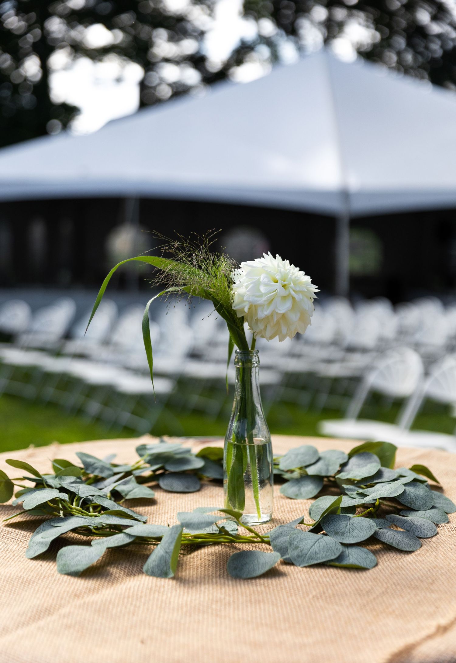 Simple wedding table centerpiece with white dahlia in glass bottle surrounded by fresh eucalyptus leaves on burlap.