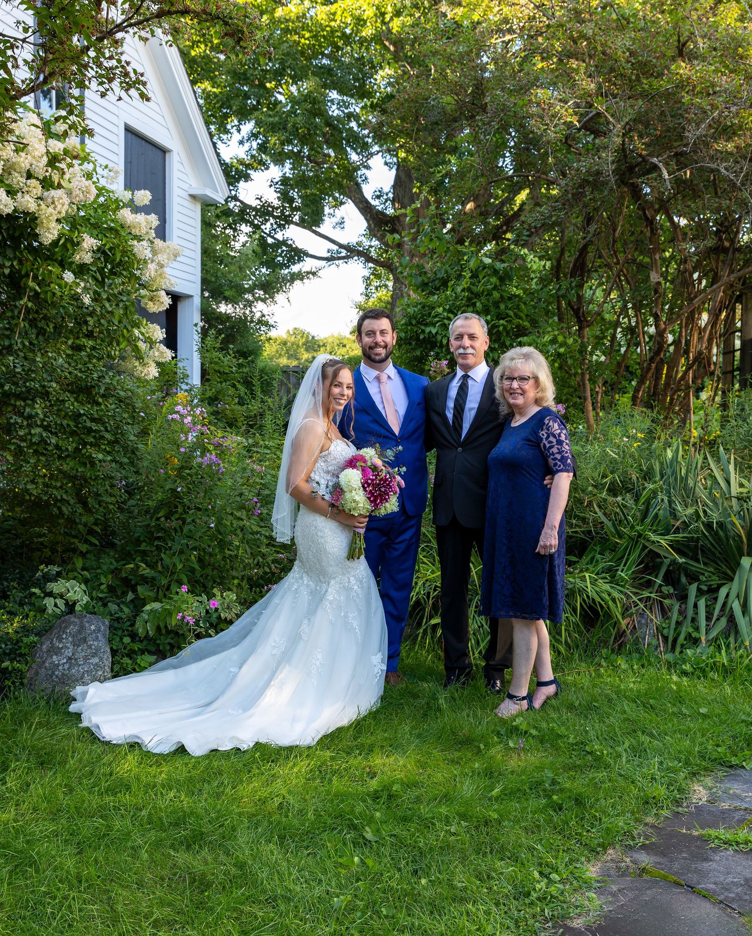 A wedding group poses in front of white house on a lush garden lawn with trees and flowers in the background.