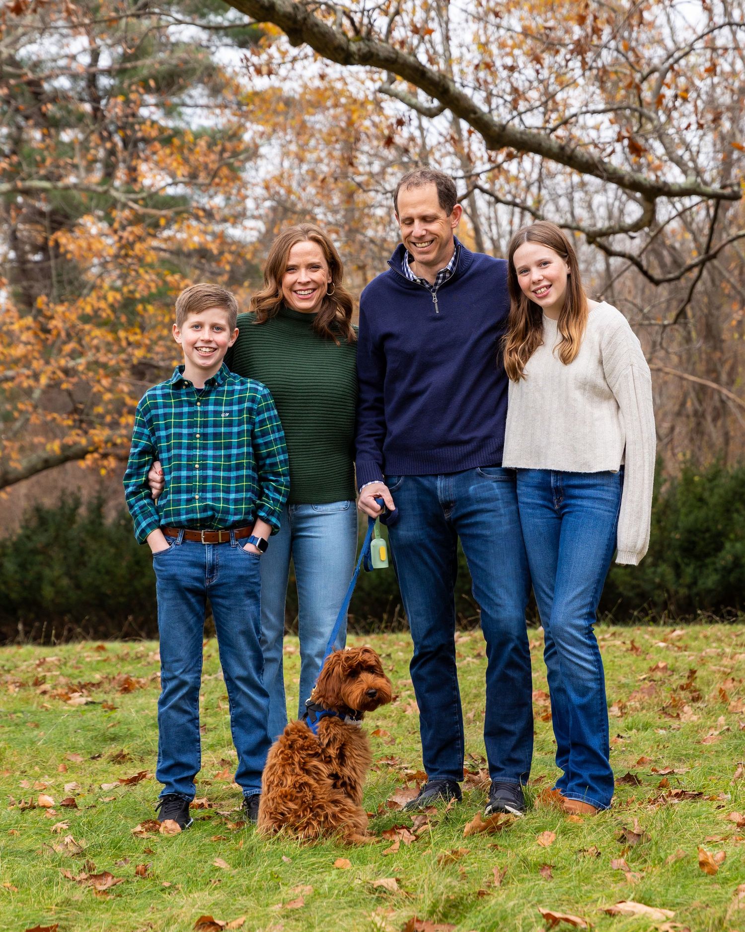 A goldendoodle dog sits in fallen autumn leaves with family members wearing coordinated blue and white outfits outdoors.