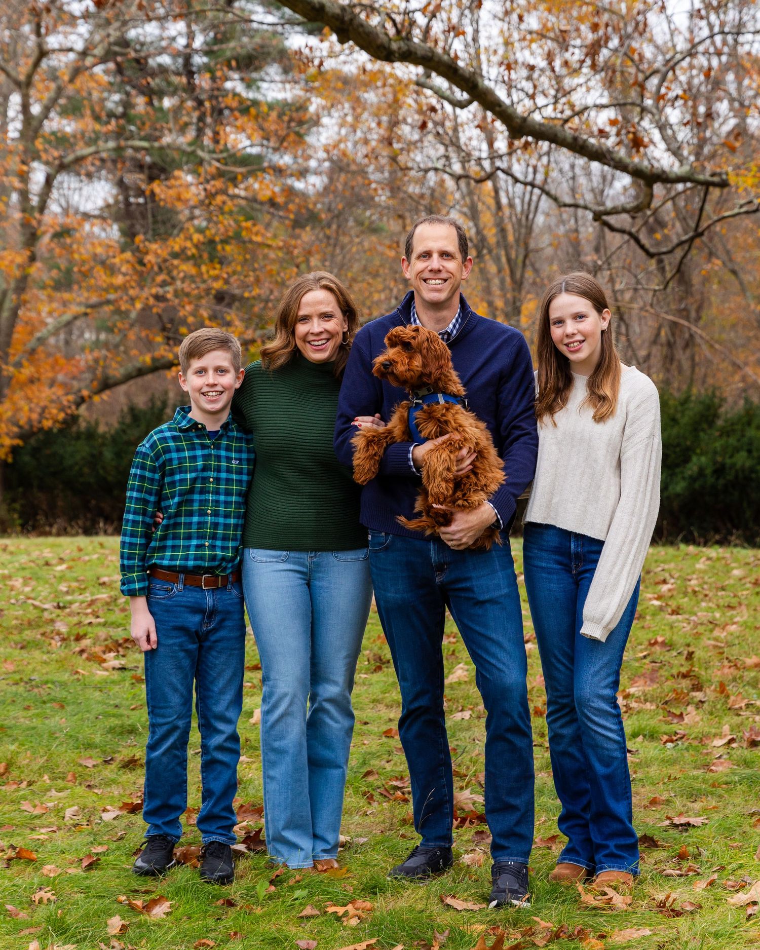 Family stands together on autumn lawn holding a brown goldendoodle dog with fall foliage in background.