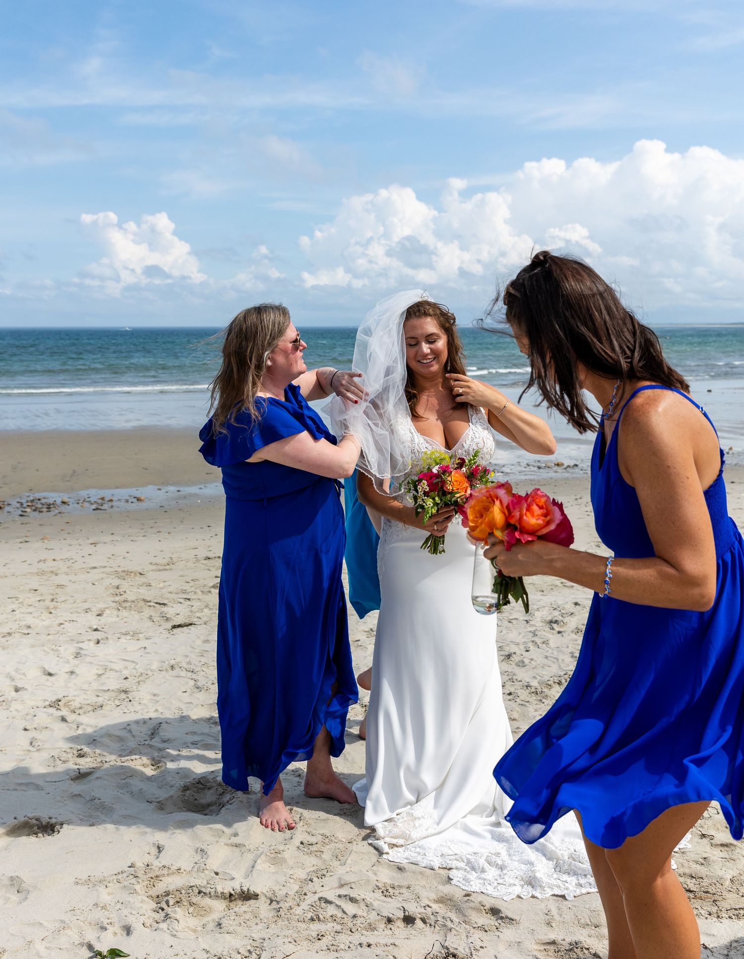 Three people in blue and white dresses share joyful moment on sunny beach with flowers and ocean backdrop.