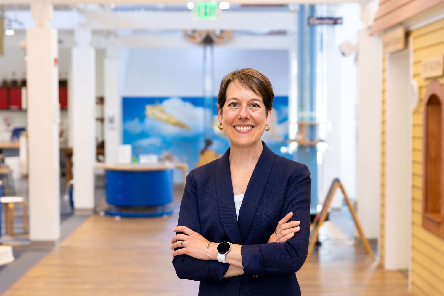 Professional in a navy blazer stands with arms crossed in a modern office space with blue accents and architectural details.