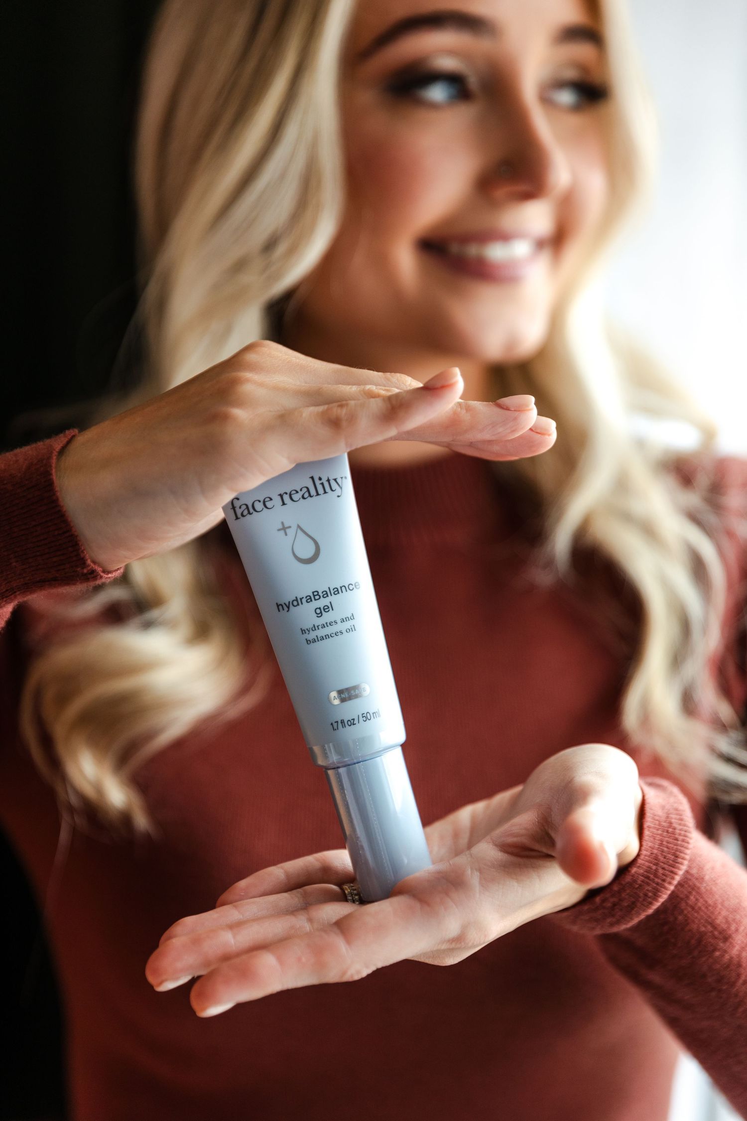 Woman in burgundy sweater holds and demonstrates a skincare product bottle against blurred background while smiling warmly.