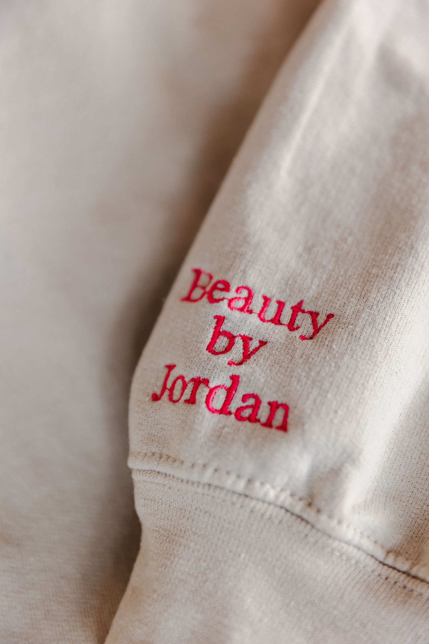 Close-up macro view of red embroidered text reading 'beauty by Jordan' on white fabric.