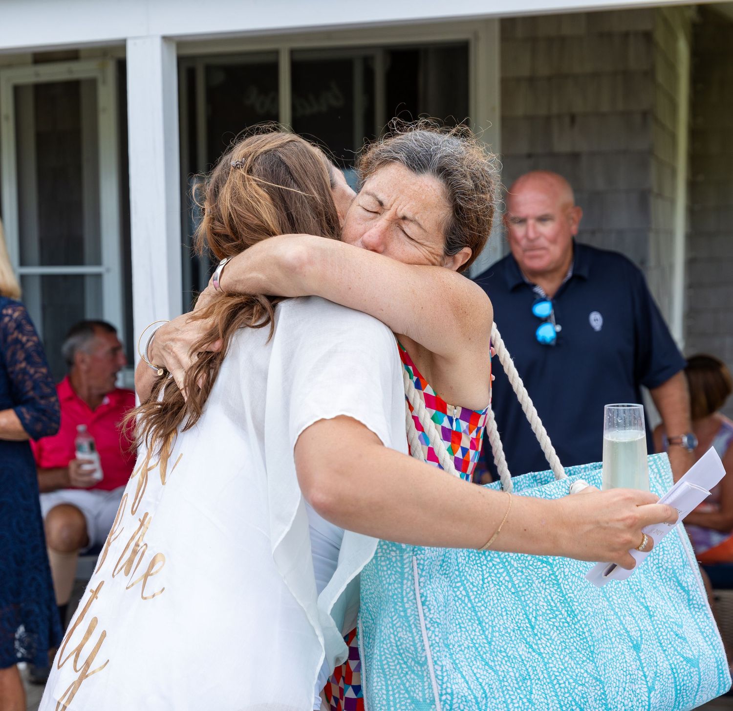 Two people embrace in an emotional hug during an outdoor gathering while others look on in the background.