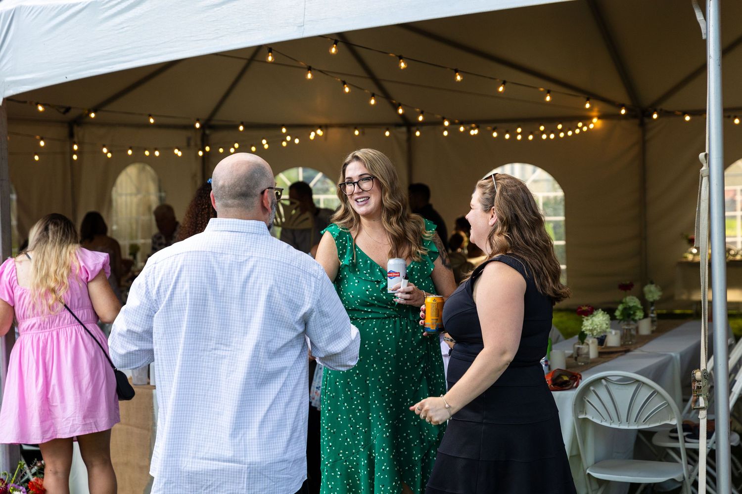 Reception guests mingle and socialize in an outdoor tent decorated with string lights and white linens.