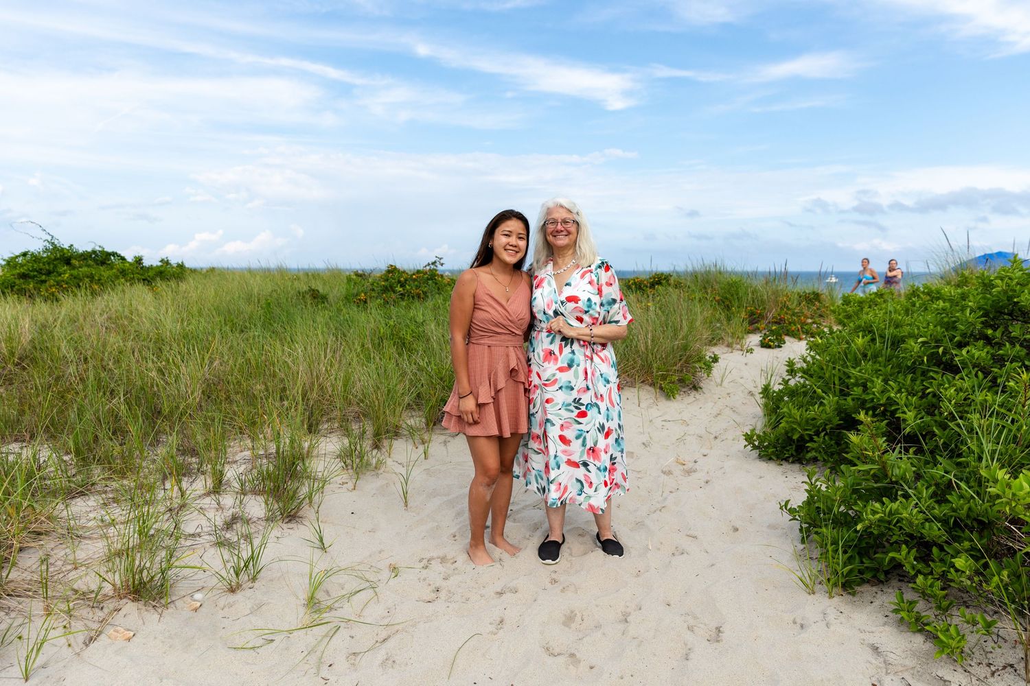 Two people walk along a sandy beach path lined with green coastal vegetation under a bright blue sky.