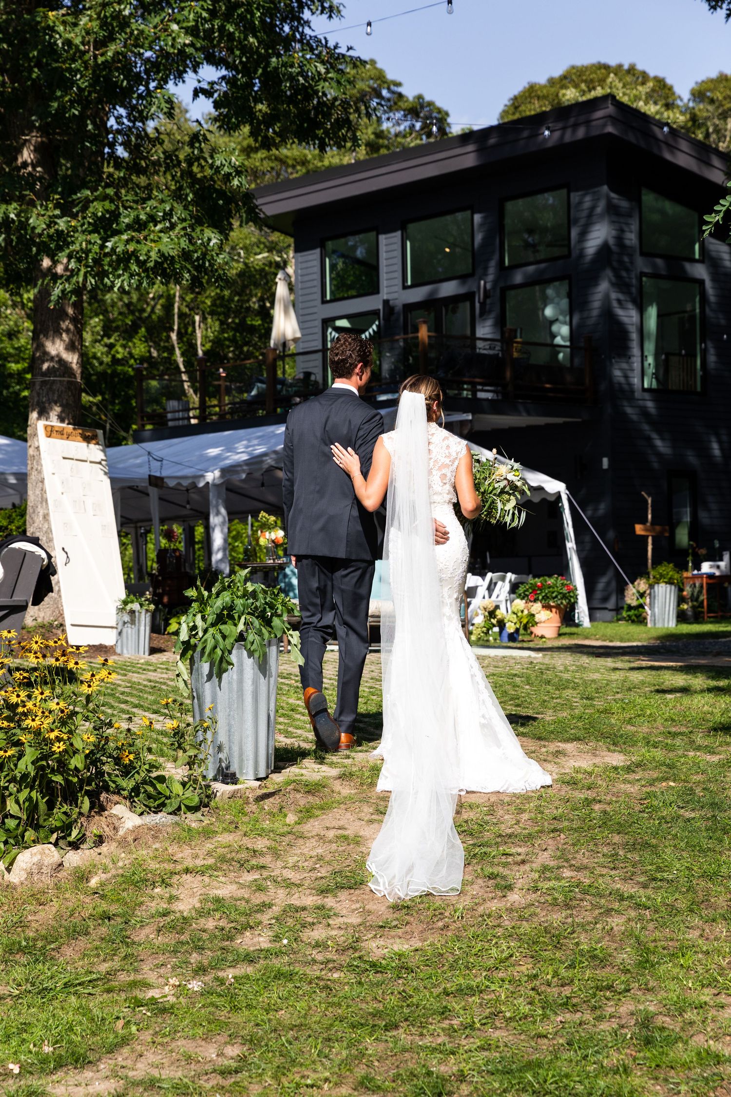 Elegant outdoor wedding ceremony setup with white decor and contemporary house in wooded background on sunny day.