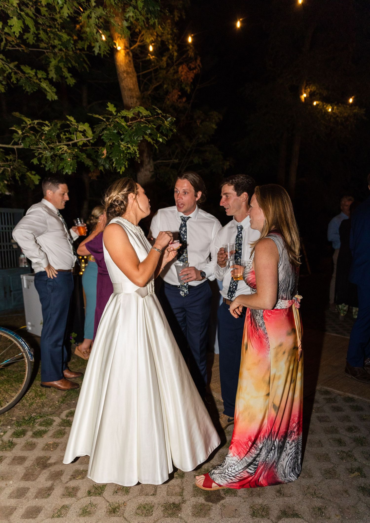 Partygoers dance and socialize at an outdoor evening celebration with string lights twinkling in the background.