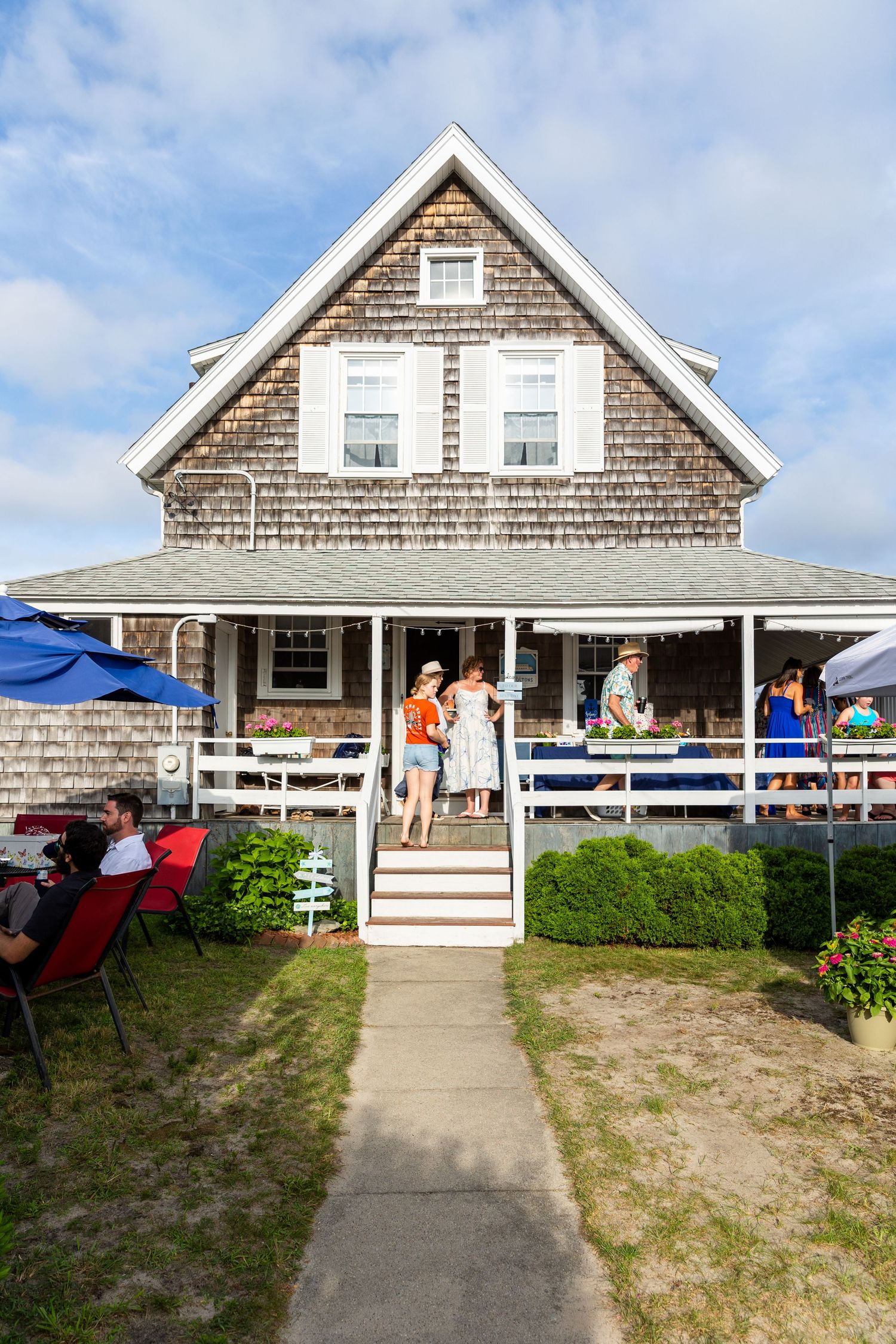 Cape Cod style beach cottage with weathered shingle siding and outdoor deck seating against blue summer sky.